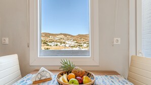 A room with a table by a window, a bowl of fruit, and a view of a coastal town. - Holiday Home 'Porto Galini Tinos Myrsini 106' with Private Terrace, Wi-Fi and Air Conditioning (LAOUTI)