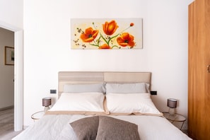 Une chambre à coucher comprenant un lit, deux tables de chevet, une commode, une armoire en bois et un tableau représentant des fleurs accroché au mur.