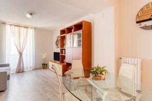 A modern living room with a glass dining table, a sofa, a wooden bookshelf, and a wall clock.