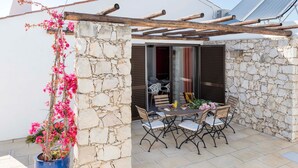 Un mur de pierre, une pergola, une table avec des chaises et un pot de fleurs orné de fleurs roses.