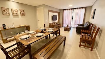 Uma sala de jantar com mesa de madeira posta para quatro lugares, um sofá e uma TV fixada na parede.