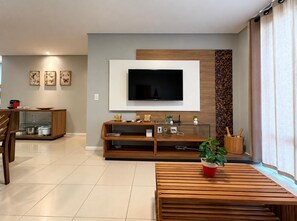 A modern living room with a flat-screen TV mounted on a wall, wooden furniture, and a dining area in the background. - GranSol, Terreo, Pé na areia, 03 qtos, Até 10 hóspedes, vizinho ao B.PARK (Aquiraz)