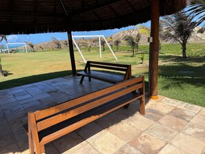 A wooden bench under a thatched roof structure with a view of a grassy area and palm trees. - GranSol, Terreo, Pé na areia, 03 qtos, Até 10 hóspedes, vizinho ao B.PARK (Aquiraz)