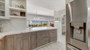 A modern kitchen with a large island, built-in cabinets, a stainless steel refrigerator, and a view of the city through the window. - Kauai Hale Awapuhi By Coldwell Banker Island Vacations (Kapaa)