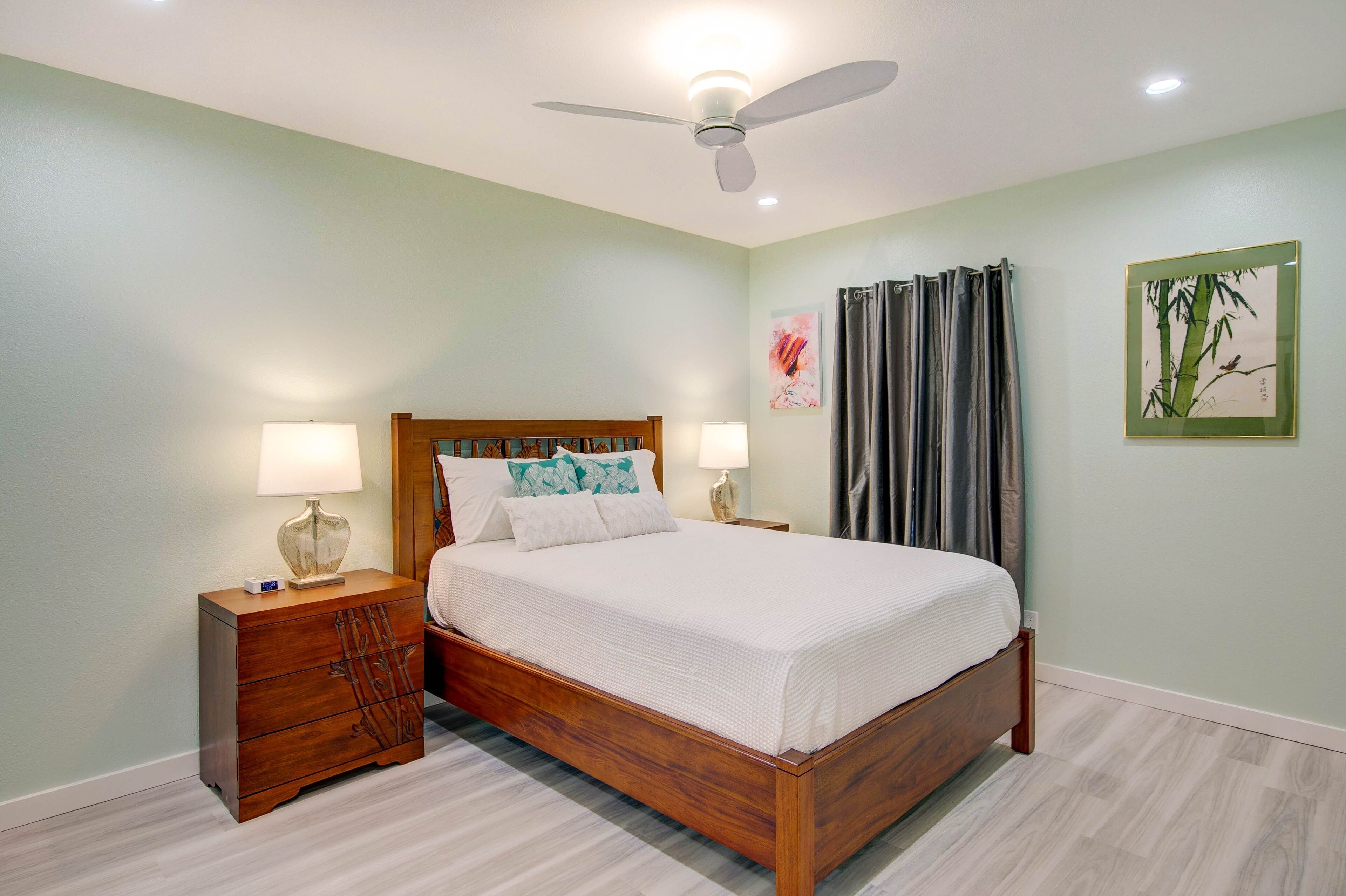 A bedroom with a wooden bed, two bedside tables with lamps, a ceiling fan, and artwork on the walls.
