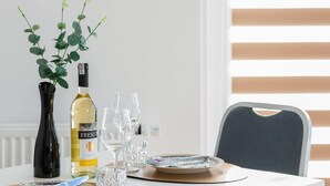 Una mesa de comedor dispuesta con una botella de vino, copas, un florero con hojas verdes y un plato con cuchara.