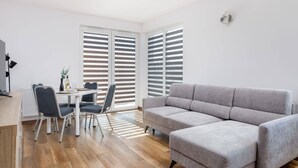 Una sala de estar moderna con un sofá gris, una mesa de comedor con sillas y ventanas grandes con persianas.