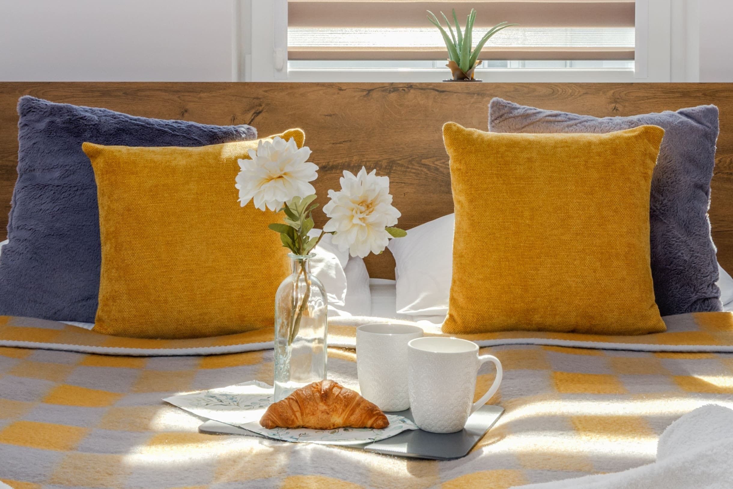 Una cama bien hecha con una colcha a cuadros, dos almohadas, un florero con flores, un croissant y dos tazas blancas.