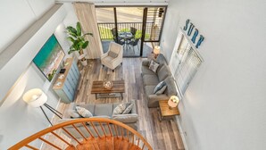 A modern interior with a spiral staircase, a living area with a sofa and coffee table, a dining table with chairs, and a large window overlooking a garden. - Kamaole Sands Two Bedrooms by Coldwell Banker Island Vacations (Kihei)