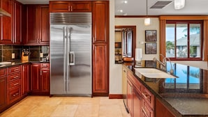 A kitchen with wooden cabinets, a stainless steel refrigerator, a large island with a sink, and a view of the outdoors through a window. - Big Island Waiulaula at Mauna Kea by Coldwell Banker Island Vacations (Kamuela)