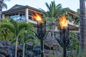 A modern hotel with a pool area, featuring two tall, black, metal torches with flames, surrounded by tropical plants and a rocky waterfall backdrop. - Ho'olei Ocean View by Coldwell Banker Island Vacations (Kihei)