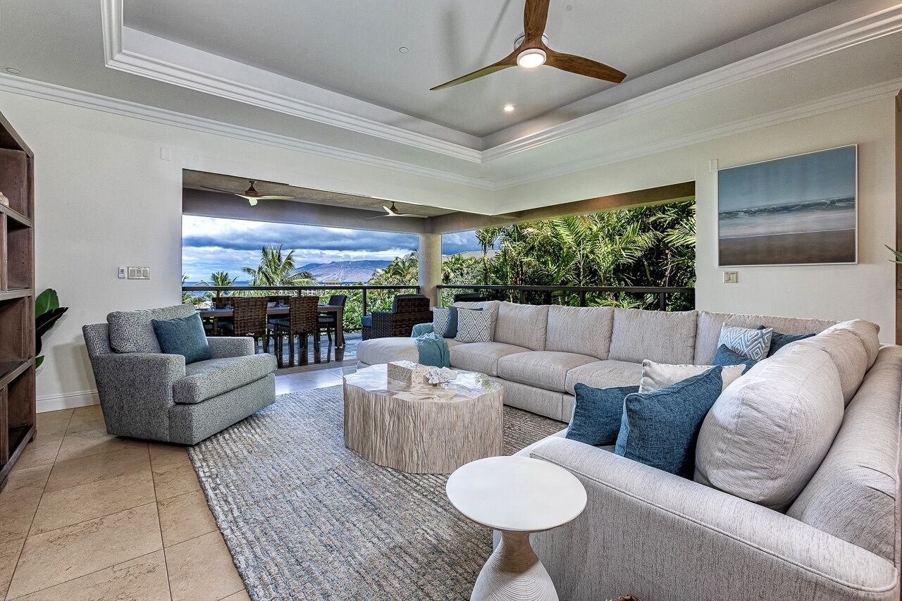 A spacious living area with a large sofa, a coffee table, and a ceiling fan. There is a view of a lush garden and a balcony with outdoor furniture.