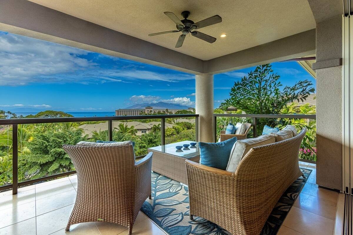 Ein Balkon mit Korbmöbeln, einem Deckenventilator und Blick auf das Meer und Gebäude.