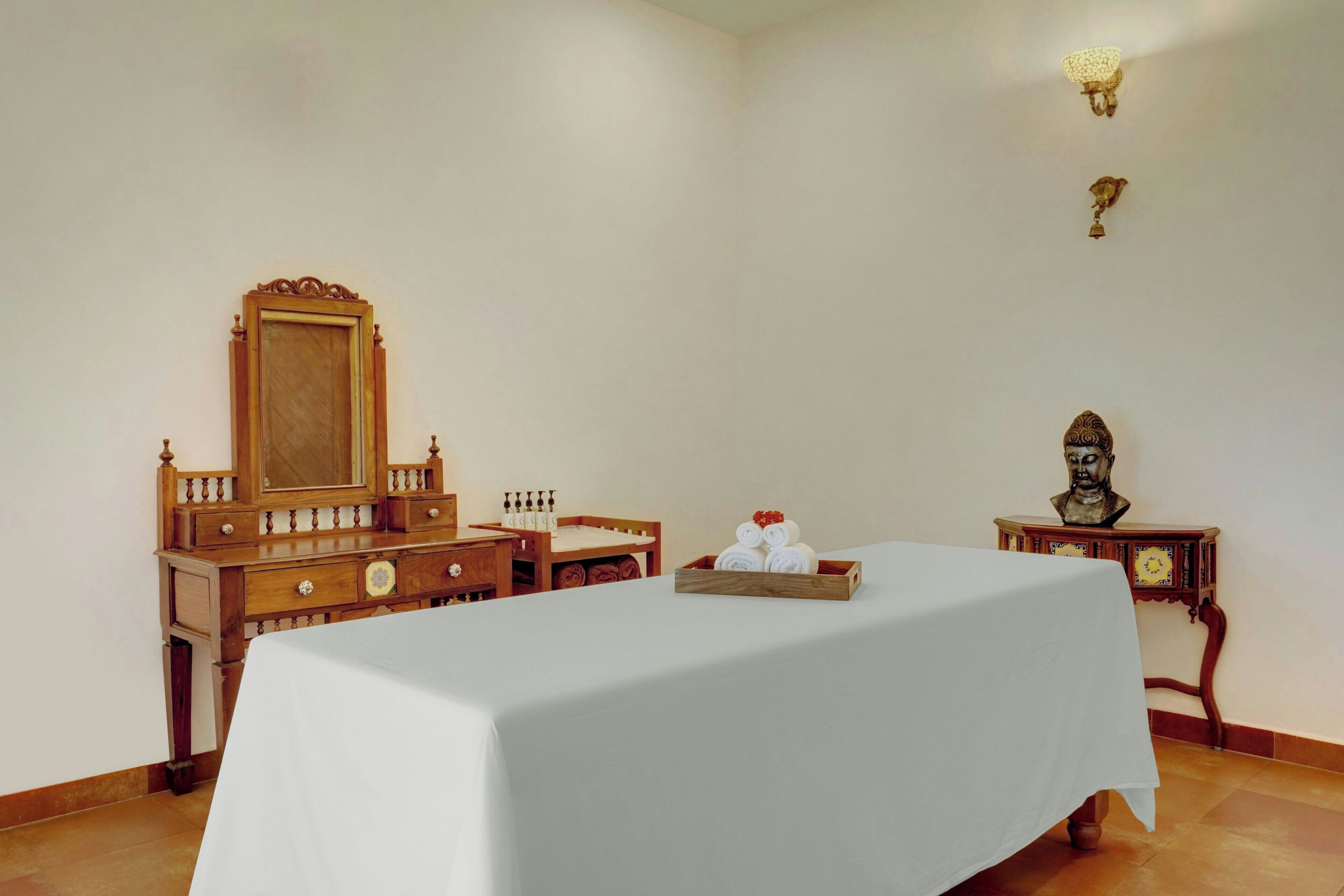 Una habitación con una cama, un tocador de madera, un espejo, un busto y un reloj sobre una mesita.