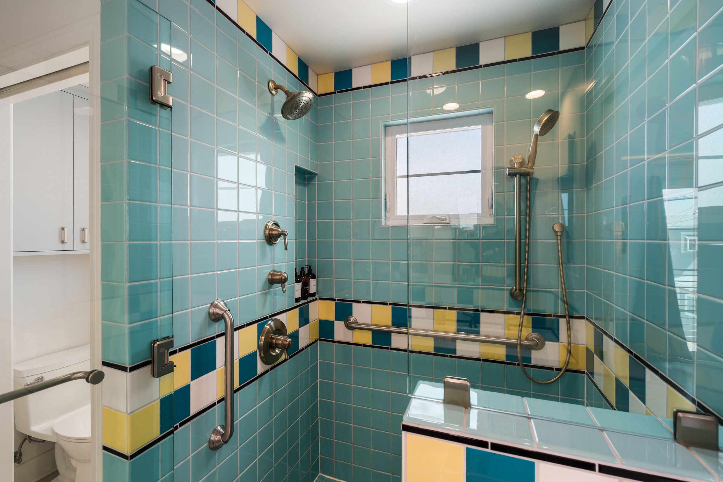 Ein Badezimmer mit blau-gelben Fliesen, einem Fenster, einer Dusche und einem WC.