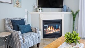 A living room with a houndstooth patterned armchair, a wooden coffee table, a fireplace with a visible fire, and a television mounted above the fireplace. - Beach Paradise VIII-DOWNTOWN WATERFRONT 3BR Luxury Condo POOL-Beach-Boardwalk (Ocean City)