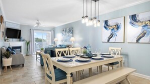 A dining area with a white table set for six, surrounded by chairs with blue cushions. There are two wall-mounted light fixtures above the table. - Beach Paradise VIII-DOWNTOWN WATERFRONT 3BR Luxury Condo POOL-Beach-Boardwalk (Ocean City)