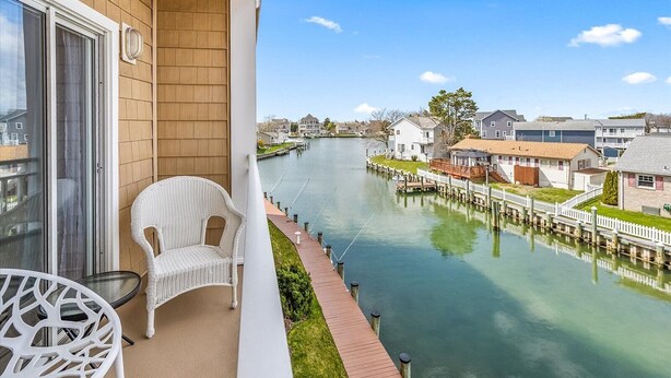 A balcony with a wicker chair and a table, overlooking a calm river with houses and docks. - Beach Paradise VIII-DOWNTOWN WATERFRONT 3BR Luxury Condo POOL-Beach-Boardwalk (Ocean City)