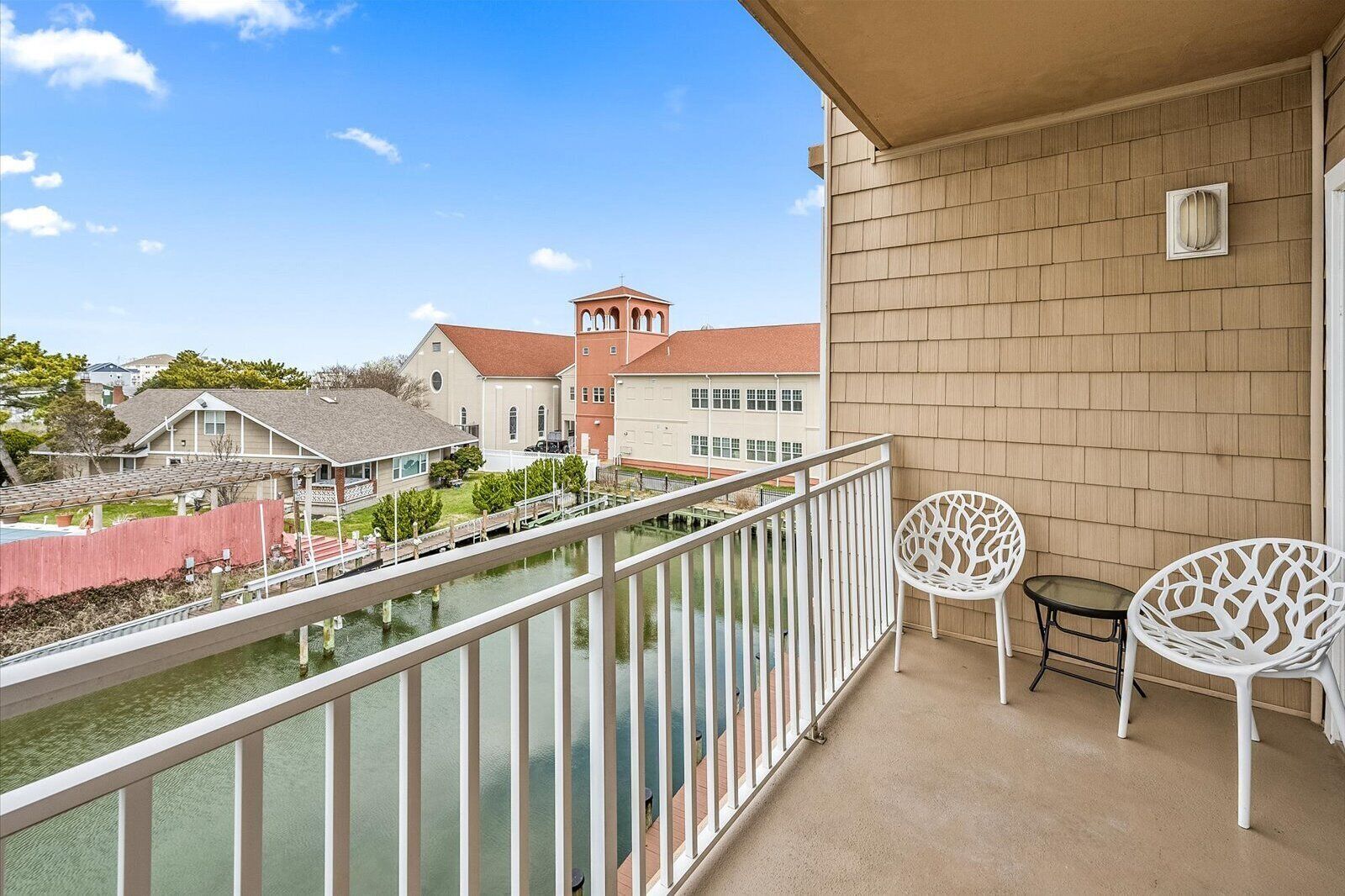 A balcony with a view of a water body and buildings. There are two white chairs and a small table.