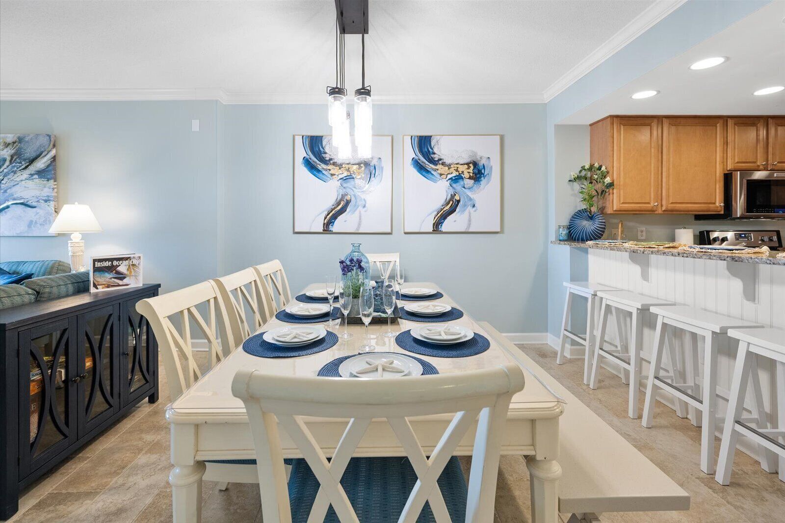 A dining area with a long table set for six, surrounded by white chairs. There are three framed abstract paintings on the wall above the table.