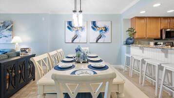 A dining area with a long table set for six, surrounded by white chairs. There are three framed abstract paintings on the wall above the table.