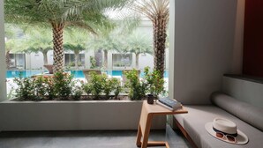 A modern room with a large window overlooking a pool and palm trees. There is a wooden side table with a book and a hat, and a cushioned bench. - Chesa Canggu (Canggu)