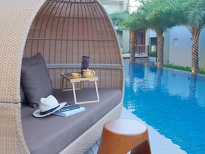 A wicker lounge chair by a pool with a table holding a laptop, a cup, and a teapot.