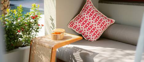 A room with a bed, a small table with a candle, a window with a view of greenery, and a patterned pillow.