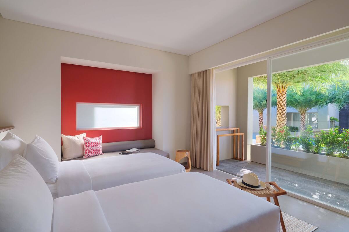 A hotel room with two beds, a red accent wall, a large window, and a view of greenery outside.