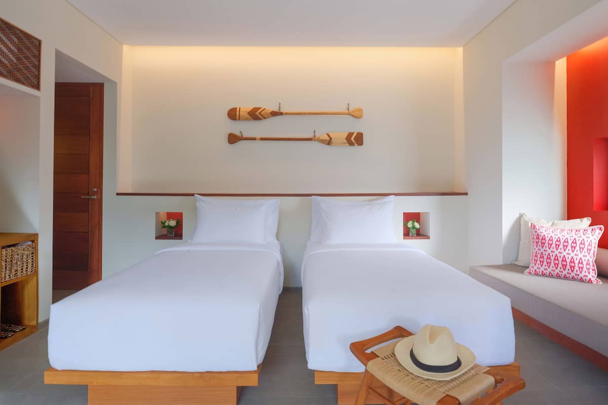 A hotel room with two beds, a wooden headboard, a wall-mounted paddleboard, and a small shelf with a vase.
