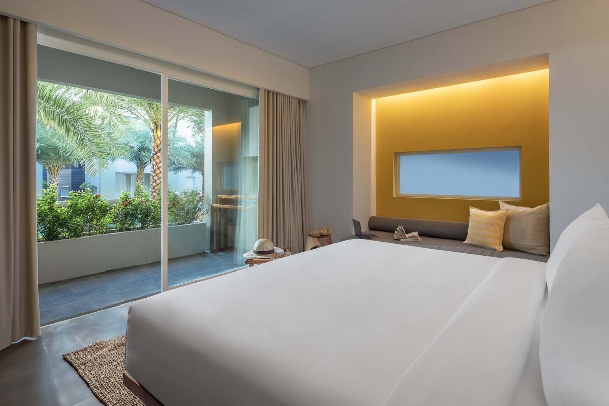 A modern hotel room with a large bed, a desk, a laptop, and a view of greenery outside.