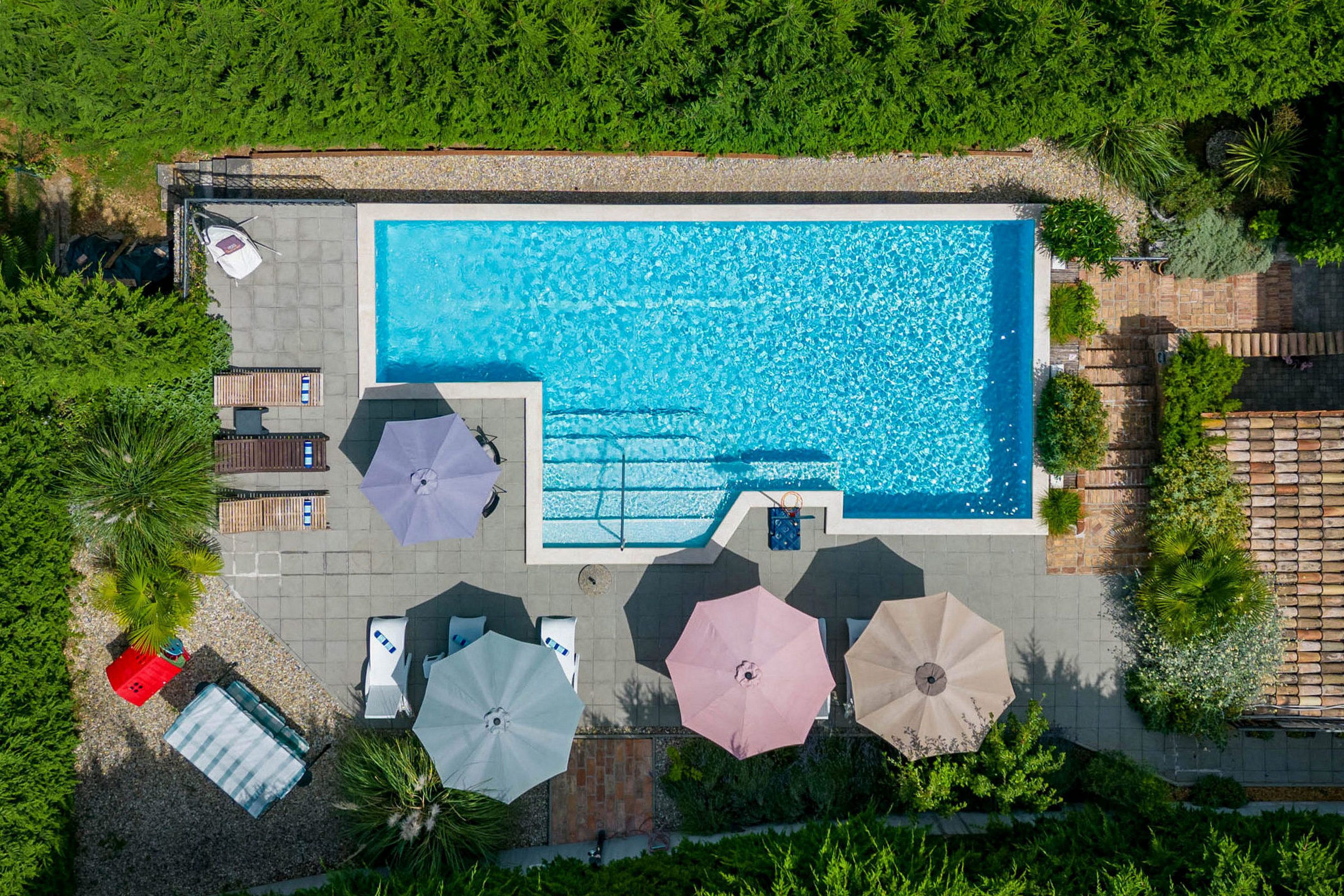 An aerial view of a rectangular swimming pool surrounded by umbrellas and lounge chairs, with a tiled deck and greenery.