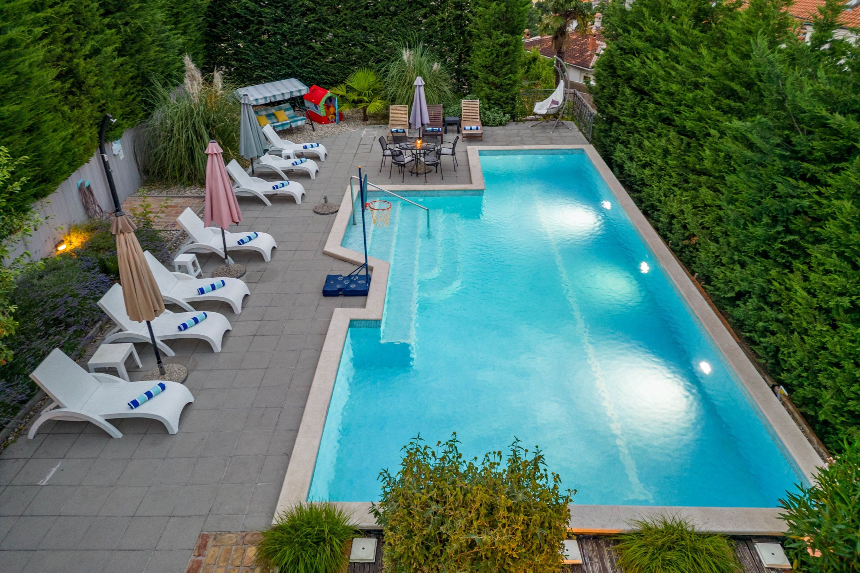 A rectangular swimming pool with a tiled deck, surrounded by lounge chairs and umbrellas.