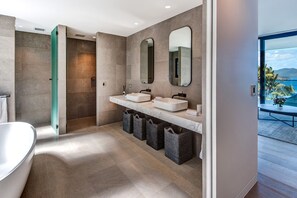 A modern bathroom with two sinks, a bathtub, and a walk-in shower. There are baskets under the sinks and a view of the outdoors through a glass door.