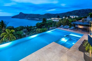 A swimming pool with a view of the sea and mountains, surrounded by lounge chairs and greenery.