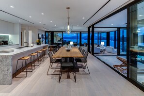 Une cuisine moderne dotée d’un comptoir en marbre et de tabourets de bar, un coin repas avec une table et des chaises en bois, et de grandes fenêtres offrant une vue sur les montagnes.