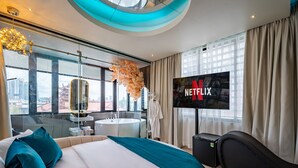 A modern hotel room with a large bed, a TV displaying the Netflix logo, a bathroom with a round bathtub, and a city view through large windows. - Midas Hotel (Ho Chi Minh City)