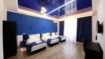 A hotel room with three beds, wooden flooring, a blue accent wall, and a ceiling light fixture.