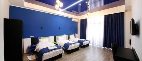 A hotel room with three beds, wooden flooring, a blue accent wall, and a ceiling light fixture.