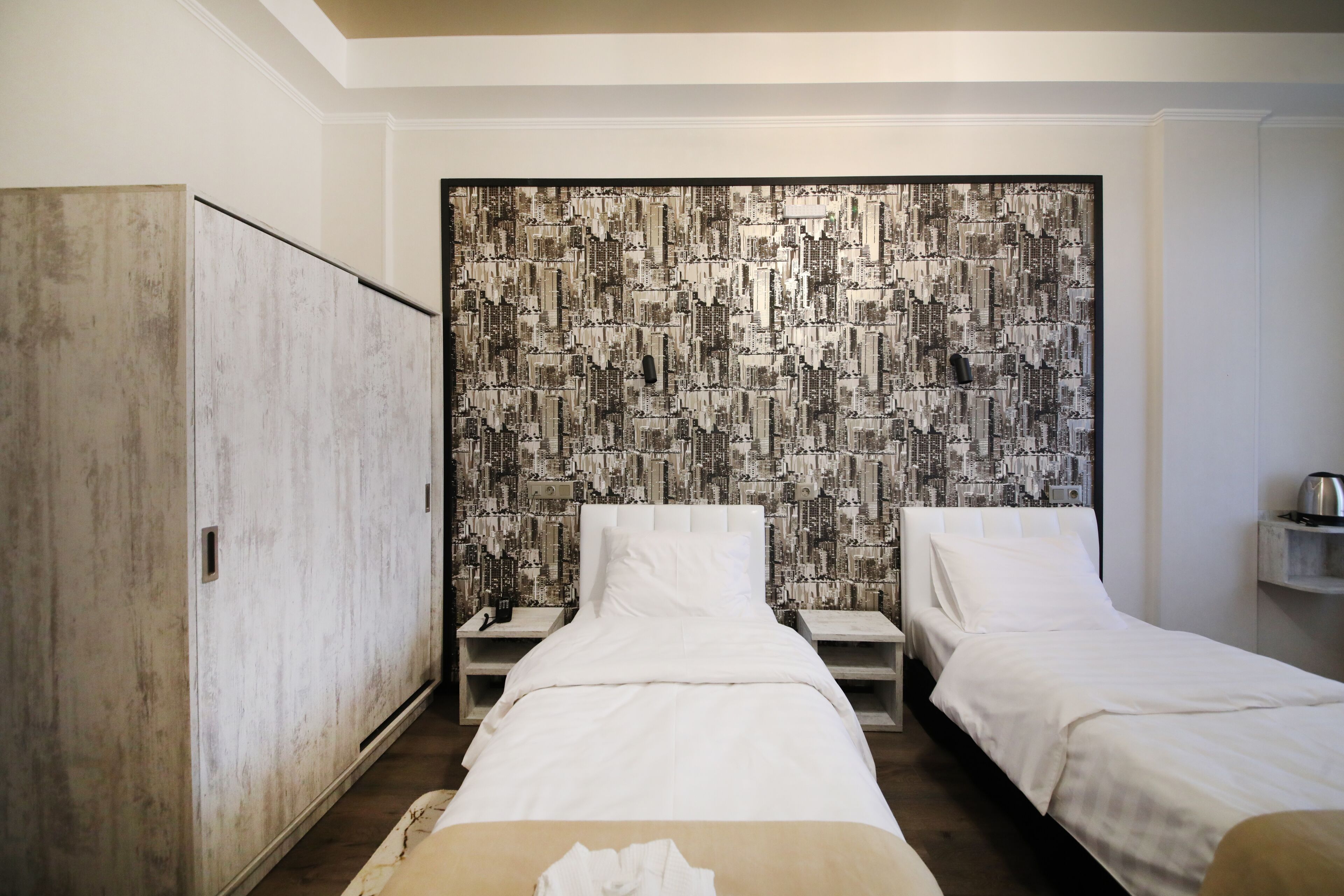 Habitación de hotel con dos camas individuales, un armario y un papel pintado con vista a la ciudad.