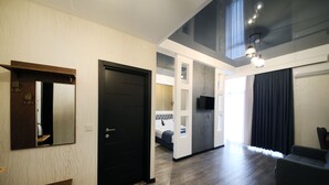 Quarto de hotel moderno com piso de madeira, porta preta, uma mesa pequena, um sofá e televisão fixada na parede.