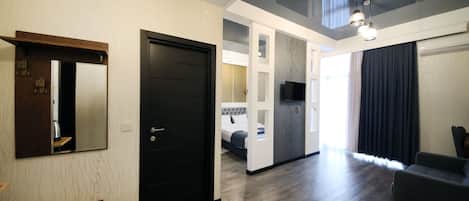 A modern hotel room with a wooden floor, a black door, a small table, a sofa, and a TV mounted on the wall.