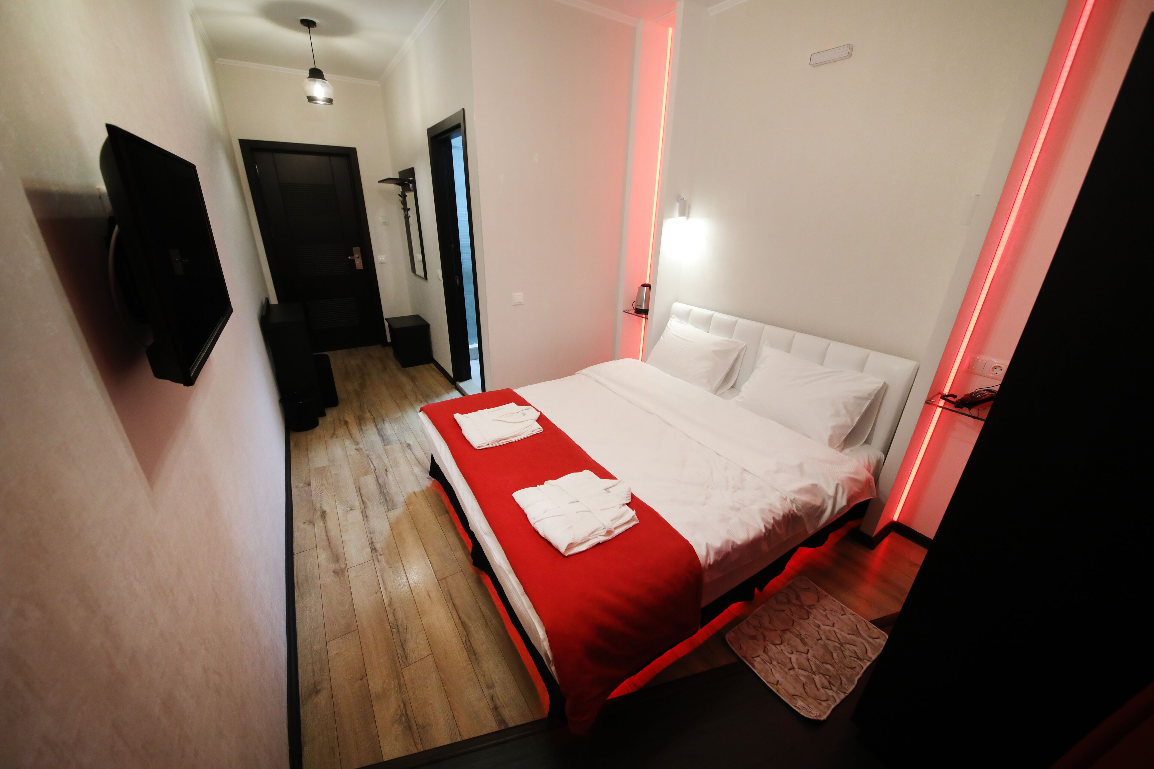 A hotel room with a bed, a TV, a red accent wall, and a hallway leading to a door.