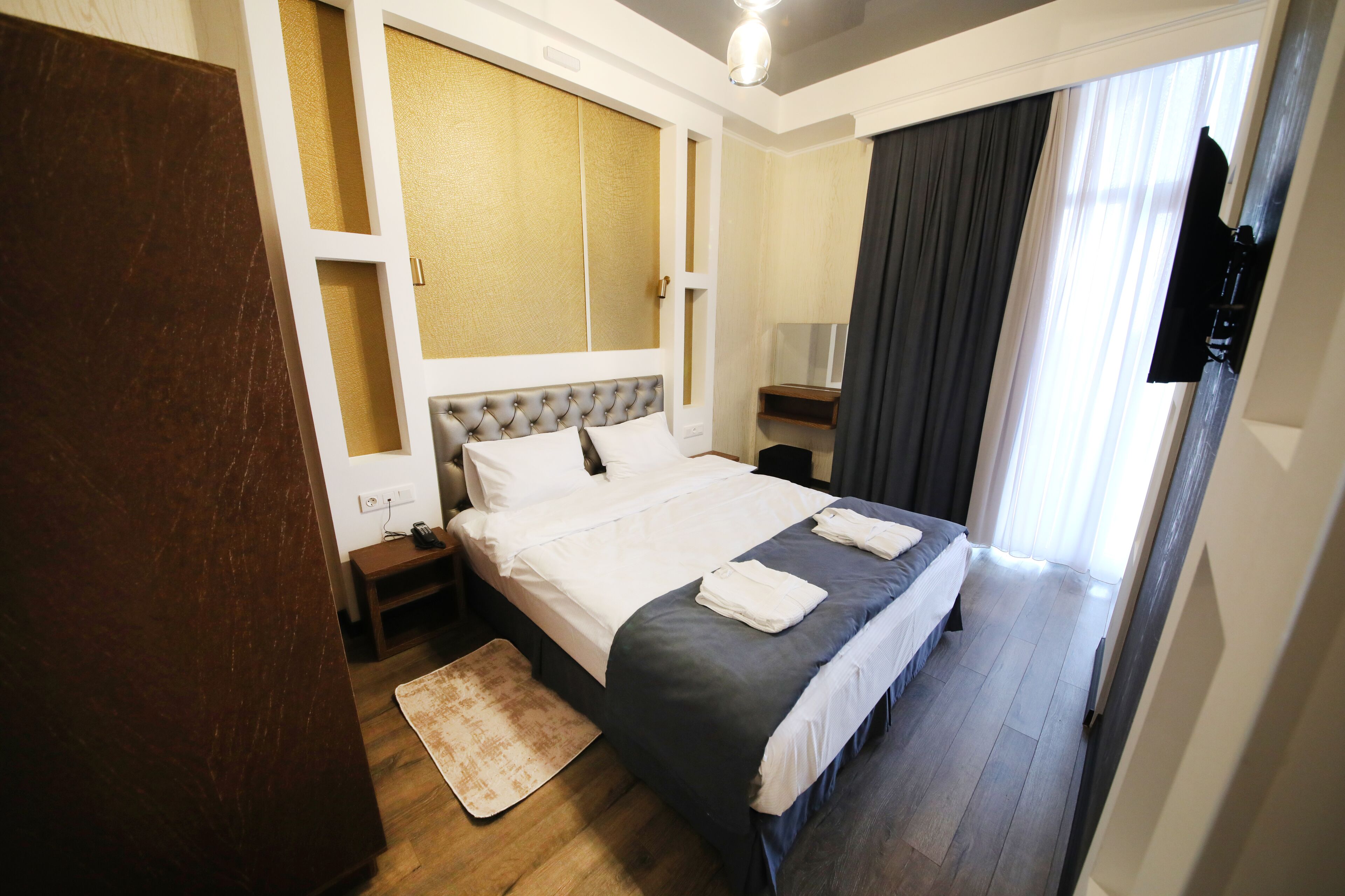 A neatly arranged hotel room with a large bed, a bedside table, a nightstand, a wall-mounted TV, and a window with curtains.
