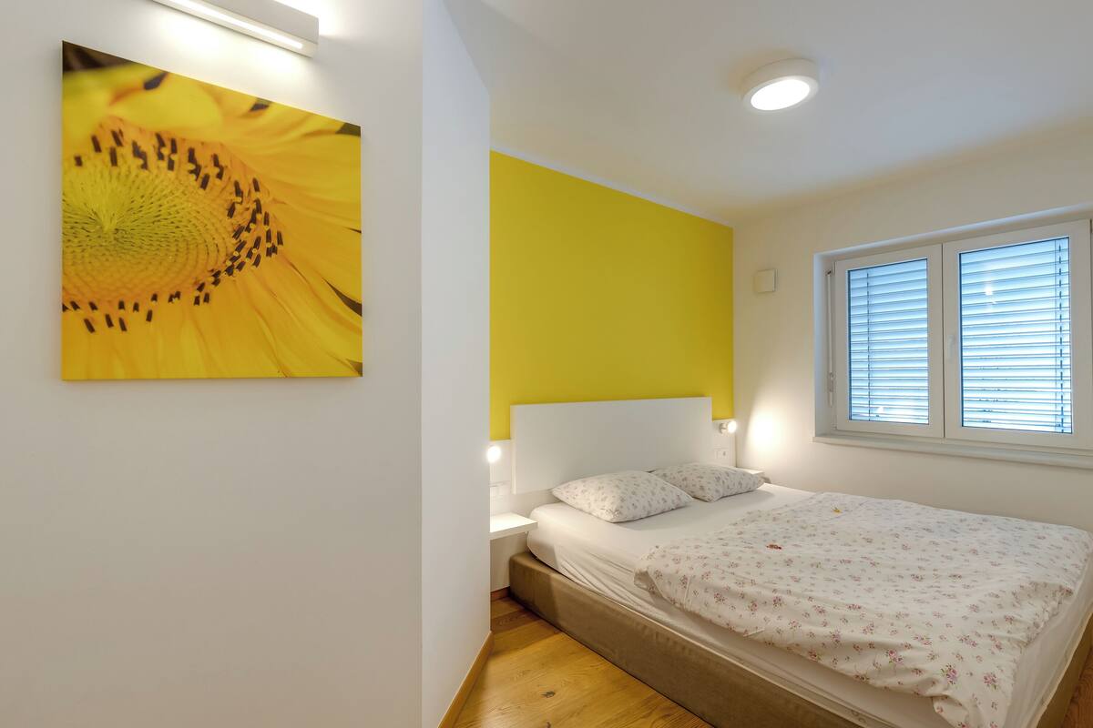 Une chambre à coucher avec un lit, une table de chevet, une lampe fixée au mur, une fenêtre avec des stores et un tableau représentant un tournesol.