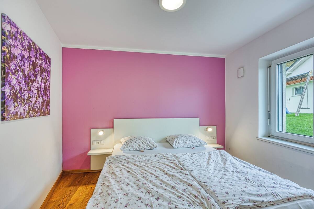 Une chambre avec un mur rose, un lit recouvert d’une literie à motifs floraux, une table de chevet et une fenêtre donnant sur une maison.