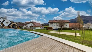 A swimming pool with a wooden deck, surrounded by houses with red roofs and a clear blue sky with clouds. - Honeycomb Chalets and Apartments Mozirje (Mozirje)