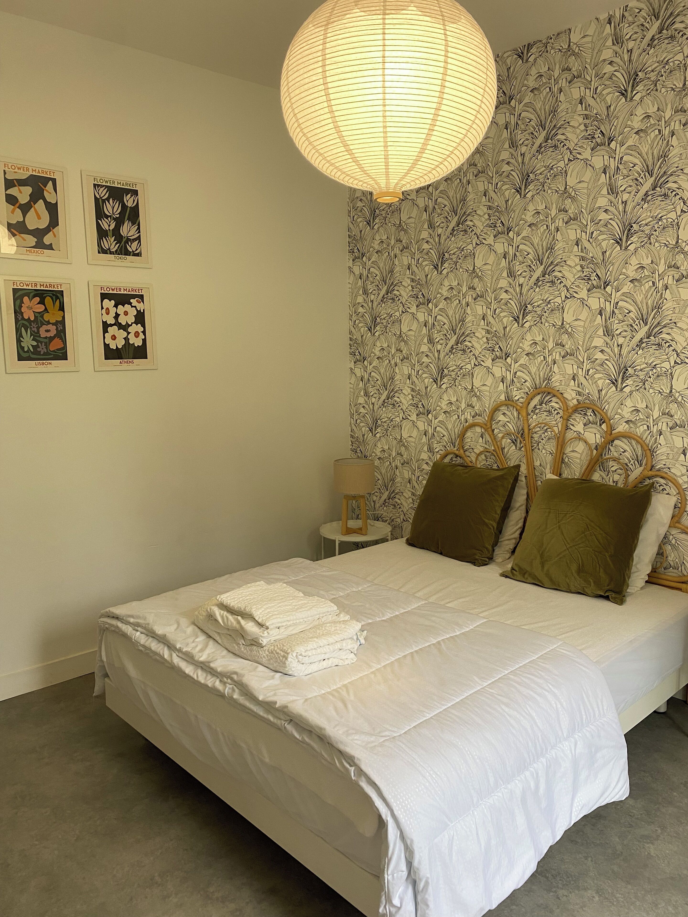 A neatly made bed with white linens, a hanging lamp, and floral wallpaper.