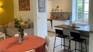 A dining area with a round table, white chairs, and a vase of flowers. There is a kitchen area with a window and a tiled backsplash. - Clos des Chardons - Le Jardin (Saint-Aignan)