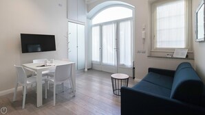 A modern living room with a flat-screen TV, a dining table with chairs, a blue sofa, and a large window with curtains. - Italianway Easy - Naviglio Pavese 6 (Milan)